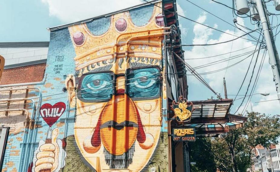 Colorful street mural of a face with a crown, holding a heart-shaped lollipop, on a blue brick wall.